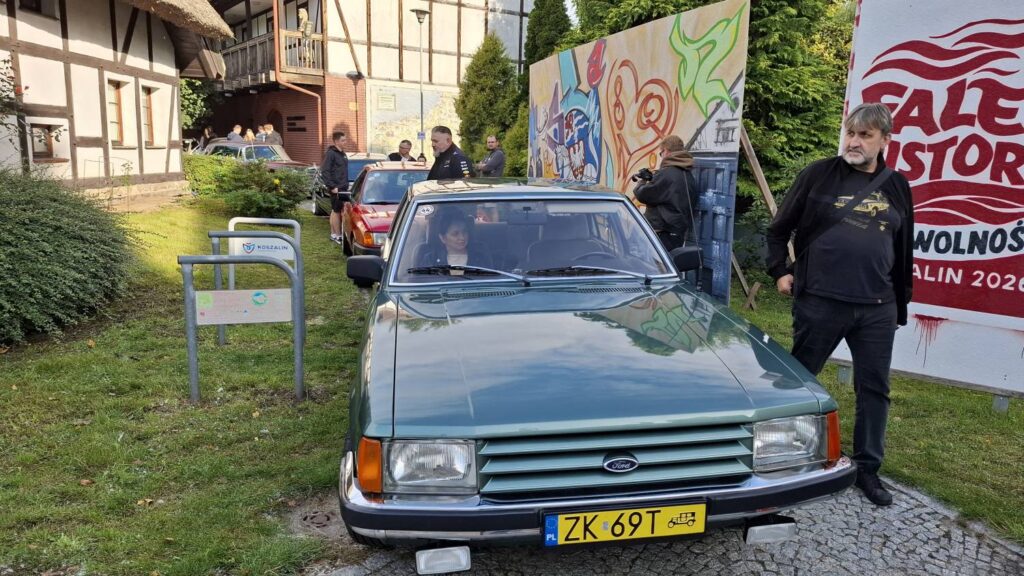 Zabytkowy samochód stoi przed muralem i banerem wydarzenia; obok pojazdu przechodzi mężczyzna, a w tle widoczne są kolejne samochody i uczestnicy zlotu (kliknięcie powiększy zdjęcie).