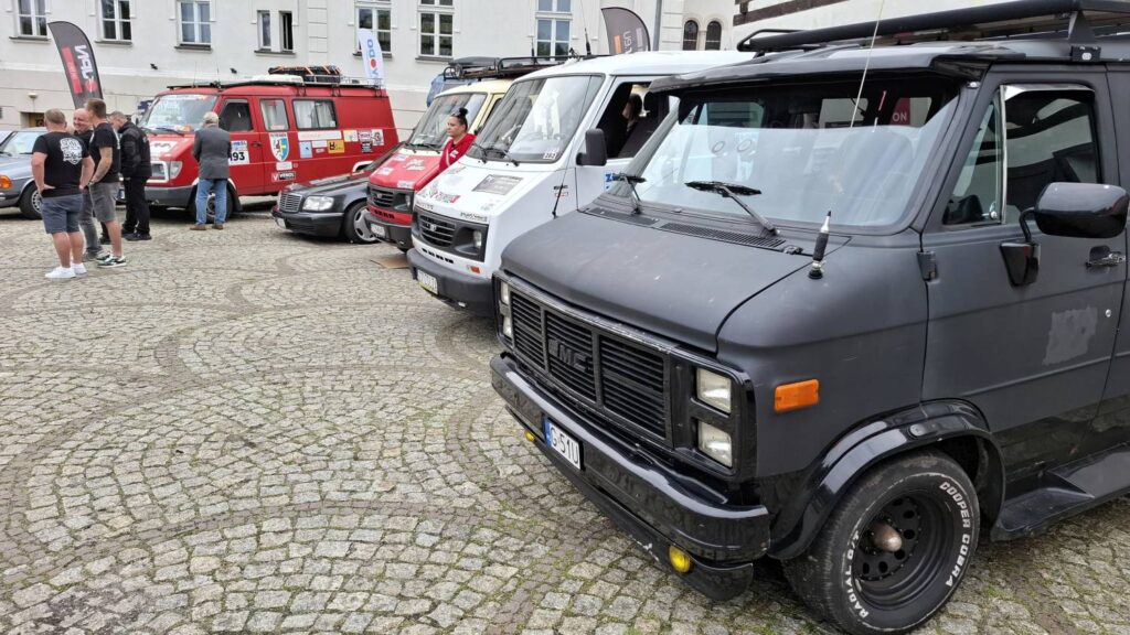 Czarny klasyczny samochód dostawczy stoi na pierwszym planie wśród innych zabytkowych pojazdów; w tle widoczne są czerwone wozy strażackie i jasne samochody dostawcze (kliknięcie powiększy zdjęcie).