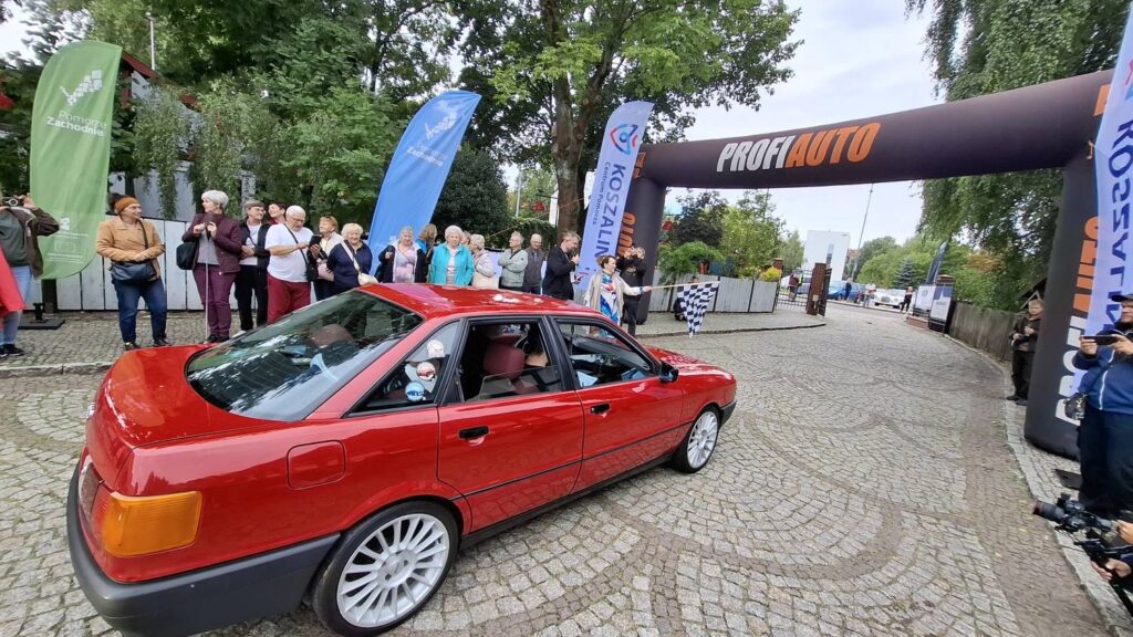 Czerwony klasyczny samochód przejeżdża przez bramę startową wydarzenia; po bokach zgromadzili się widzowie, a nad trasą widoczny jest łuk z napisem „PROFIAUTO” (kliknięcie powiększy zdjęcie).
