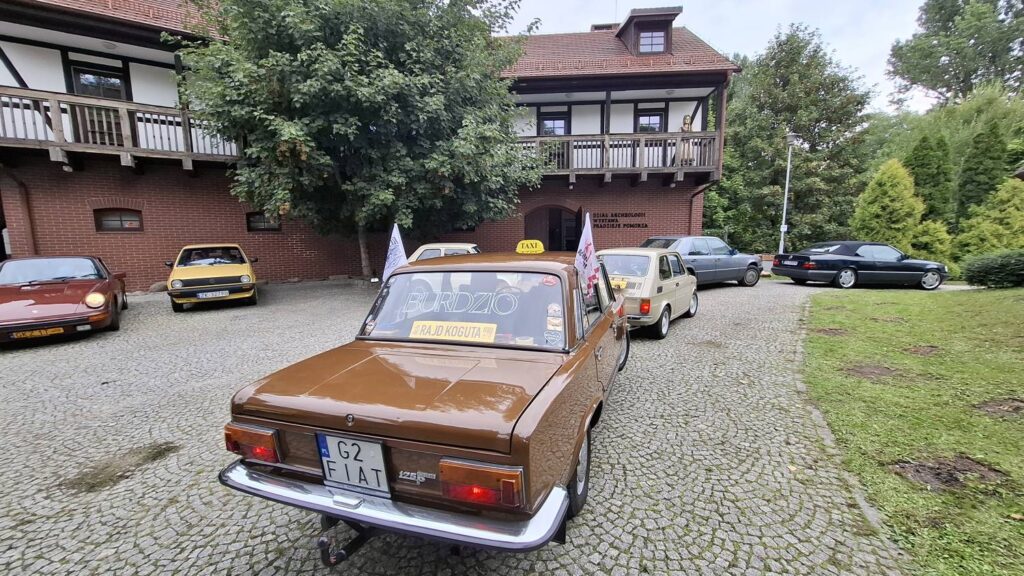 Brązowy klasyczny samochód z napisem „FIAT” na tylnej szybie stoi na brukowanym dziedzińcu; w tle zaparkowane są żółte, białe, srebrne i czarne samochody, a za nimi znajduje się drewniana zabudowa (kliknięcie powiększy zdjęcie).