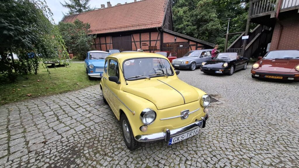 Żółty klasyczny Fiat 500 stoi na pierwszym planie na brukowanym dziedzińcu; w tle widoczne są niebieskie, czerwone, szare i czarne samochody oraz zabytkowy budynek drewniany (kliknięcie powiększy zdjęcie).