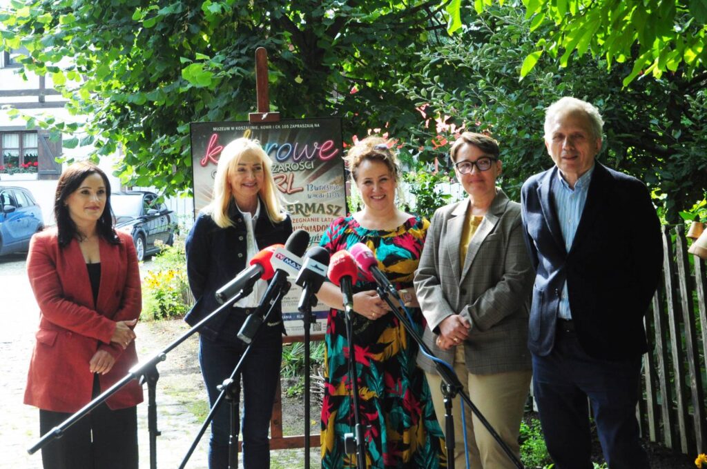 Uczestnicy konferencji prasowej stoją przed mikrofonami na zewnątrz; w tle widoczny jest plakat wydarzenia oraz zielona roślinność (kliknięcie powiększy zdjęcie).