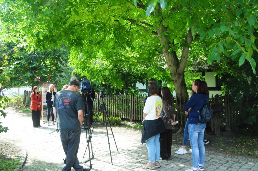 Ekipa lokalnej telewizji rejestruje konferencję prasową na świeżym powietrzu; operator kamery nagrywa osoby występujące przed mikrofonami, a pozostali uczestnicy obserwują wydarzenie (kliknięcie powiększy zdjęcie).