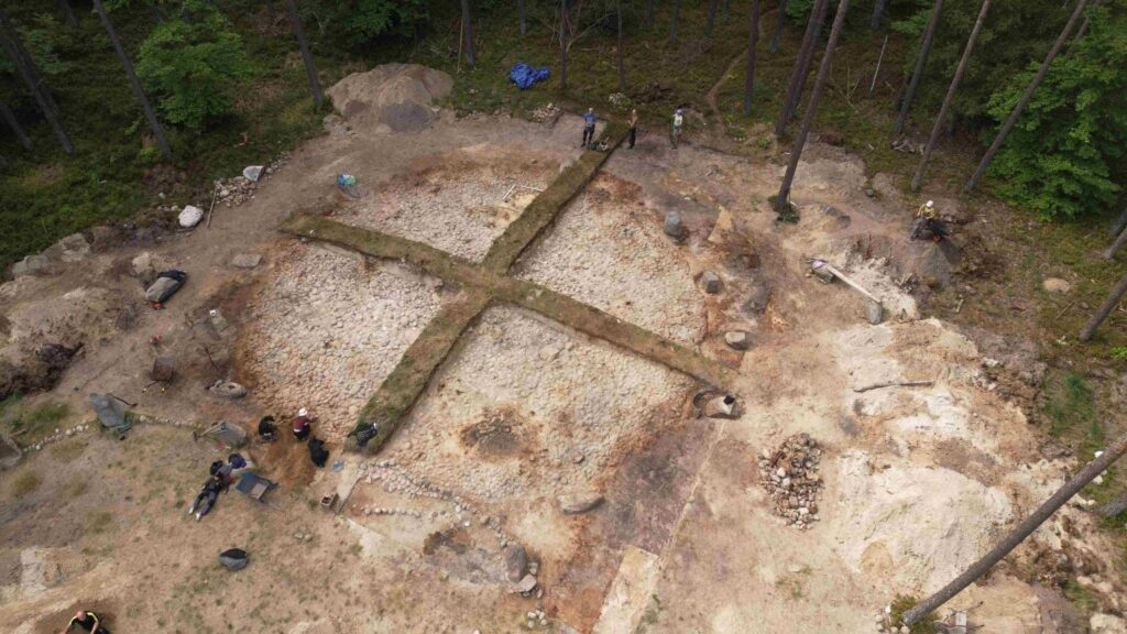 Zdjęcie wykonane z lotu ptaka przedstawiające rozległe stanowisko archeologiczne wśród lasu; widoczne są wytyczone obszary wykopalisk, odsłonięte struktury i pracujący badacze (kliknięcie powiększy zdjęcie).