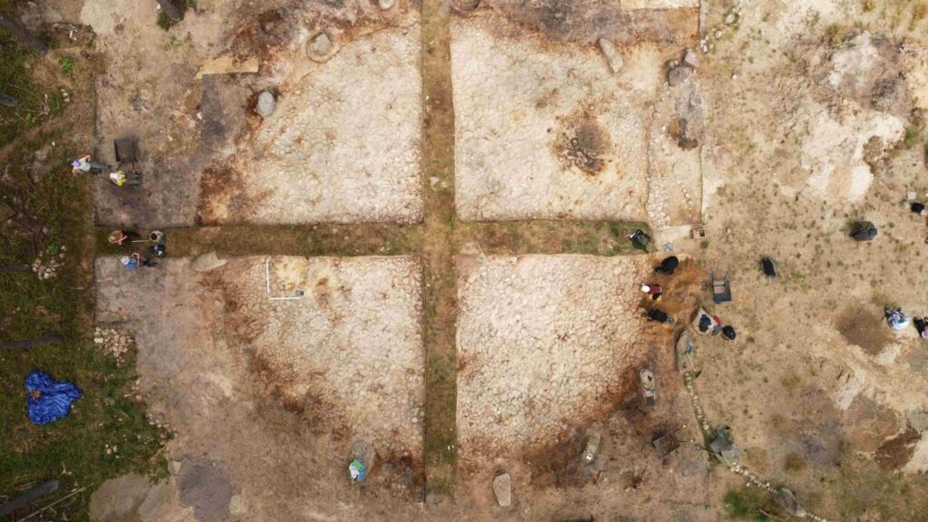 Pionowe ujęcie z lotu ptaka ukazujące fragment stanowiska archeologicznego podzielony na kwatery badawcze; na odsłoniętej powierzchni widoczne są ślady wykopów, kamienie oraz osoby prowadzące prace (kliknięcie powiększy zdjęcie).