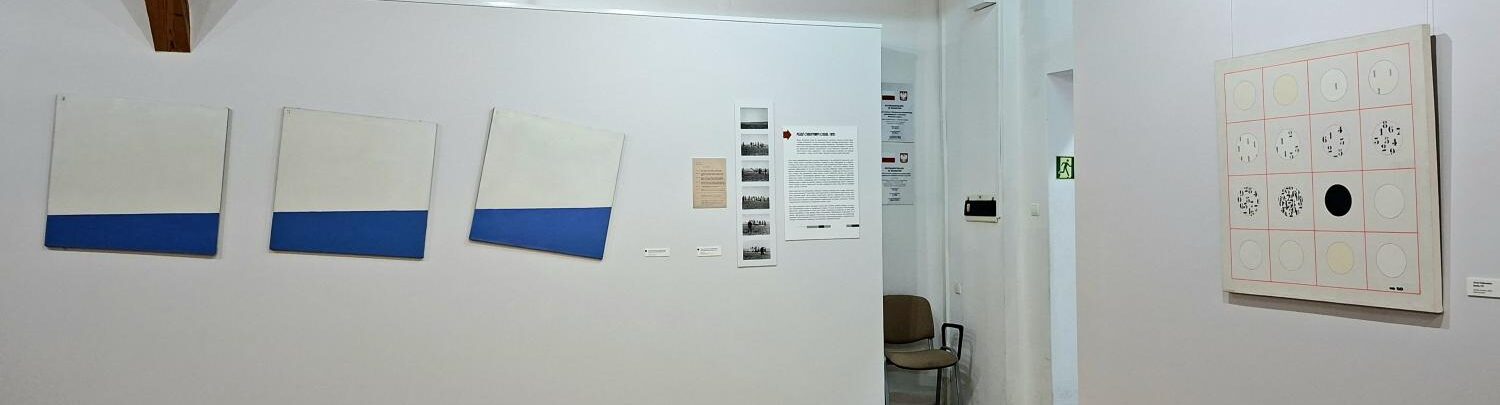 Widok fragmentu wystawy z minimalistycznymi obrazami w odcieniach bieli i granatu oraz grafiką przedstawiającą układ okręgów na przeciwległej ścianie (kliknięcie powiększy zdjęcie).
