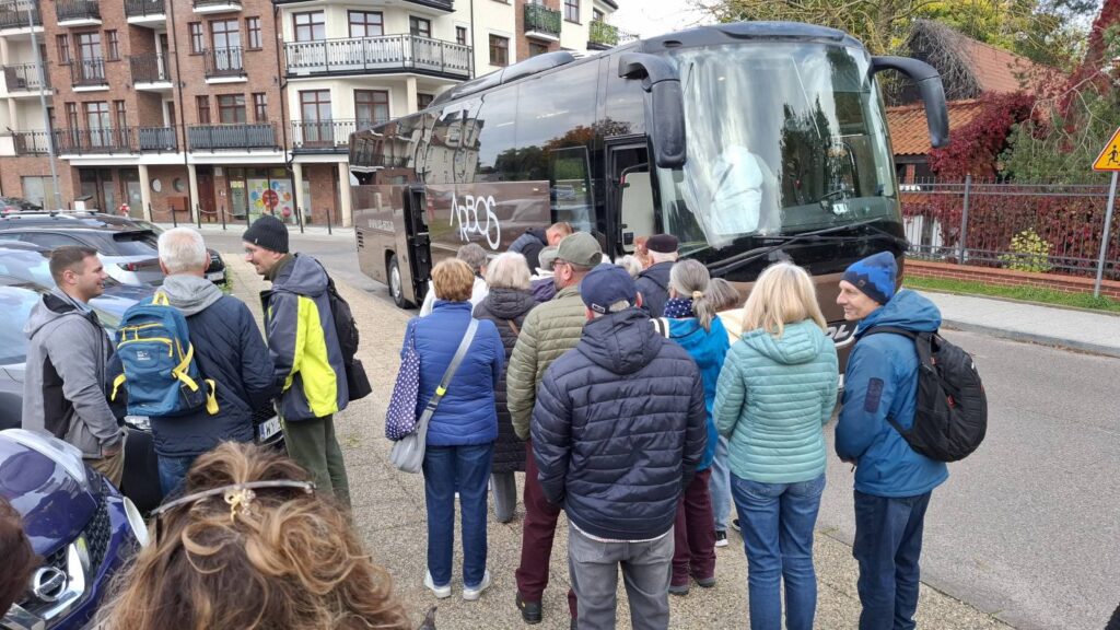 Grupa uczestników stoi obok autokaru zaparkowanego przy ulicy; osoby z plecakami i torbami przygotowują się do wejścia na pokład (kliknięcie powiększy zdjęcie).