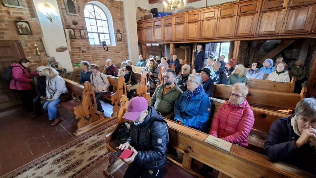 Grupa zwiedzających siedzi w drewnianych ławkach w zabytkowym kościele; wnętrze zdobią drewniane empory, obrazy i dekoracje religijne, a część osób trzyma ulotki lub notatki (kliknięcie powiększy zdjęcie).