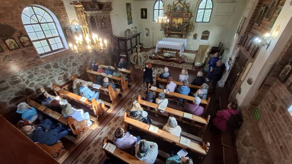 Widok z góry na wnętrze zabytkowego kościoła; widoczne są rzędy drewnianych ławek, kamienne ściany, żyrandole oraz ołtarz, przy którym zgromadzili się uczestnicy wycieczki (kliknięcie powiększy zdjęcie).