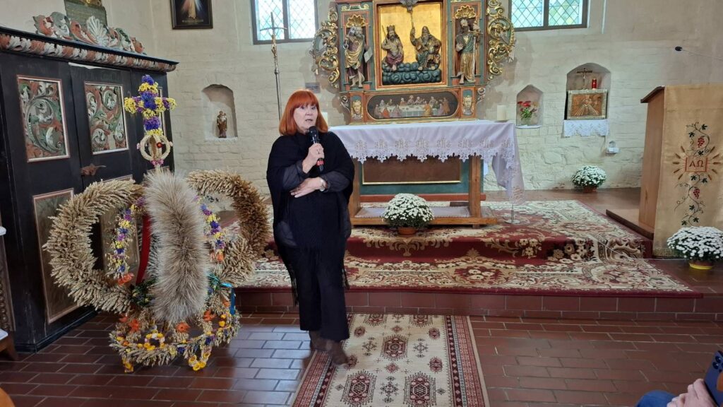 Kobieta przemawia przed grupą w prezbiterium zabytkowego kościoła; za nią znajduje się ołtarz z obrazami i dekoracjami, a po lewej stronie stoją wieńce z suszonych roślin (kliknięcie powiększy zdjęcie).