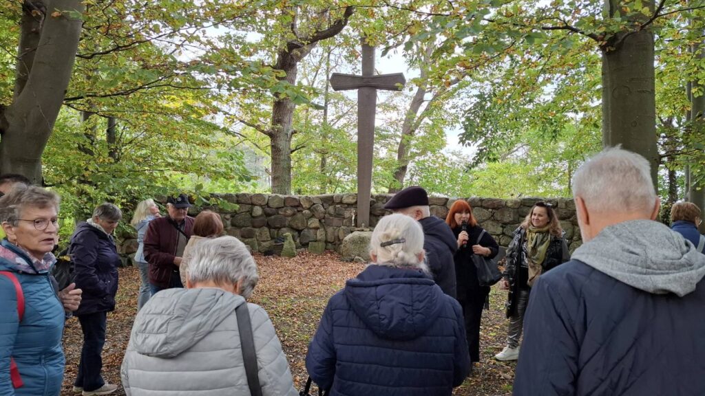 Grupa uczestników wycieczki stoi wśród drzew przy kamiennym murze i dużym kamiennym krzyżu; osoby słuchają oprowadzania w jesiennym otoczeniu (kliknięcie powiększy zdjęcie).