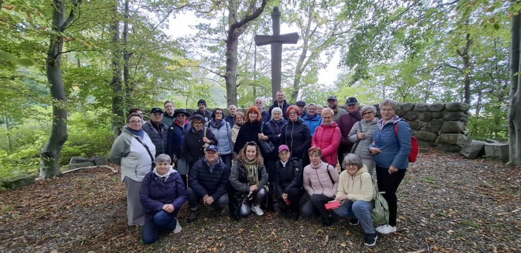 Uczestnicy wycieczki pozują do pamiątkowego, grupowe zdjęcia na tle kamiennego muru i krzyża; w tle drzewa i krzewy miejscowego parku (kliknięcie powiększy zdjęcie).