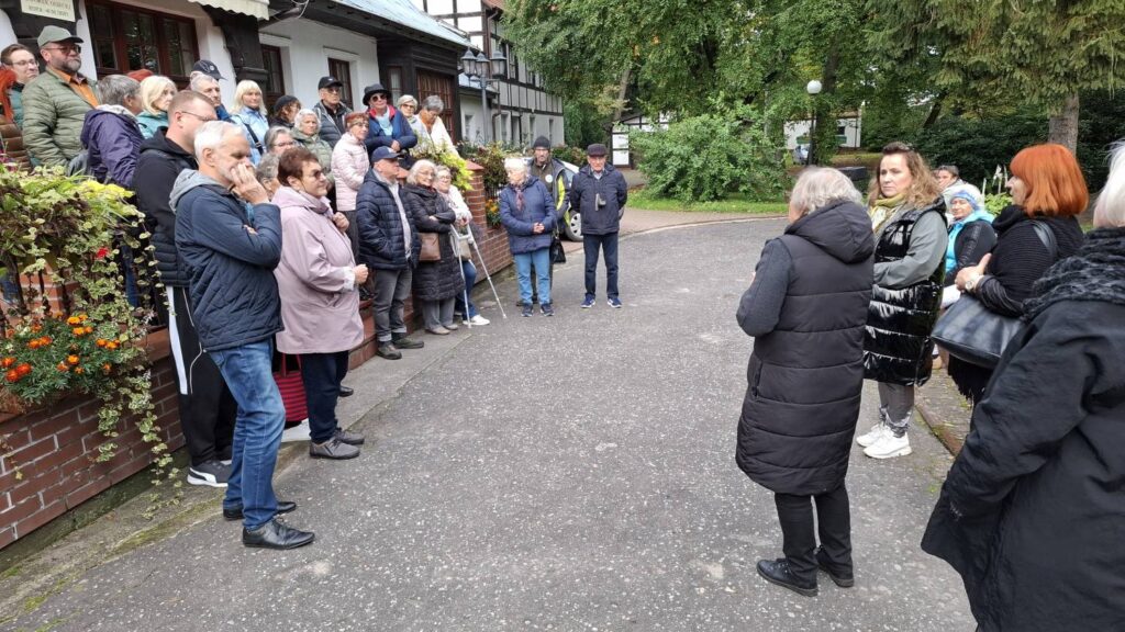 Uczestnicy wycieczki stoją przed budynkiem w otoczeniu zieleni; przewodnik opowiada o obiekcie, a grupa słucha na dziedzińcu przy ozdobnych rabatach kwiatowych (kliknięcie powiększy zdjęcie).