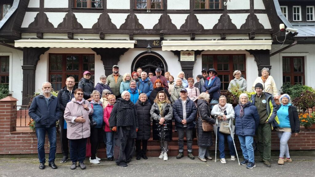 Grupa uczestników wycieczki pozuje do wspólnego zdjęcia przed wejściem do budynku o konstrukcji szachulcowej; za grupą widoczne są drewniane drzwi, okna i dekoracyjne elementy elewacji (kliknięcie powiększy zdjęcie).