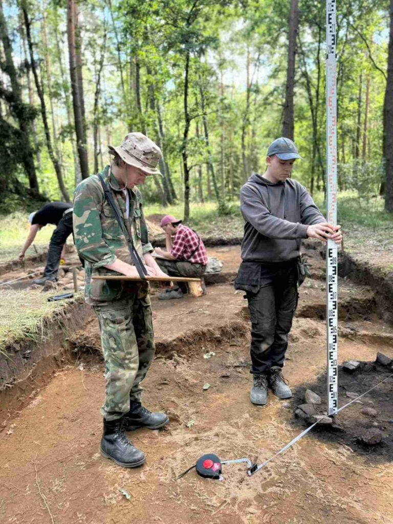 Dwóch mężczyzn wykonuje pomiary archeologiczne w wykopie na leśnym stanowisku; jeden w ubraniu moro zapisuje wyniki w notesie, drugi w szarej bluzie ustawia pionową łatę pomiarową, w tle pozostali uczestnicy pracują przy ziemi (kliknięcie powiększy zdjęcie).