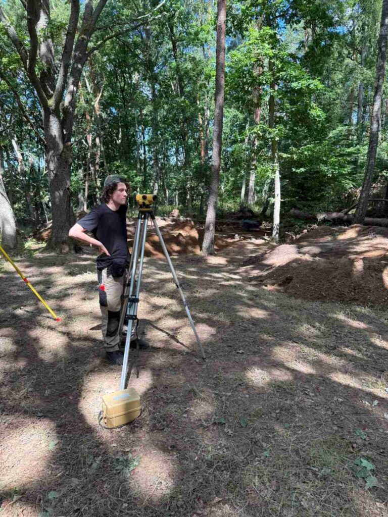 Mężczyzna obsługuje instrument pomiarowy na statywie podczas badań archeologicznych w lesie; obok urządzenia leży żółta walizka ze sprzętem, a w tle widoczne są wykopy i hałdy ziemi między drzewami (kliknięcie powiększy zdjęcie).