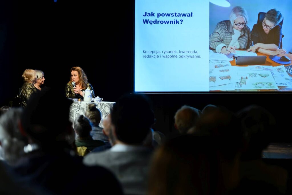 Trzy kobiety rozmawiają na scenie obok stolika z białym obrusem; na dużym ekranie widoczny jest slajd „Jak powstawał Wędrownik?” z fotografią osób pracujących nad publikacją, a na pierwszym planie widać publiczność (kliknięcie powiększy zdjęcie).