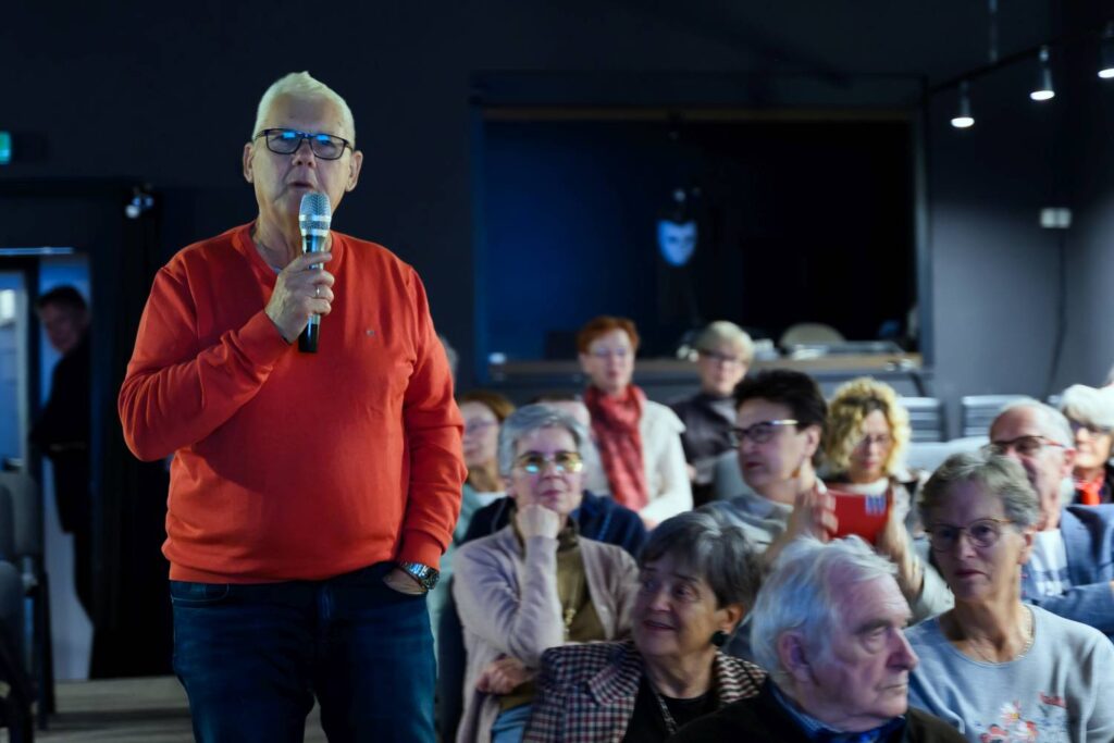 Siwowłosy mężczyzna w czerwonym swetrze i okularach trzyma mikrofon i wypowiada się na tle siedzącej publiczności (kliknięcie powiększy zdjęcie).