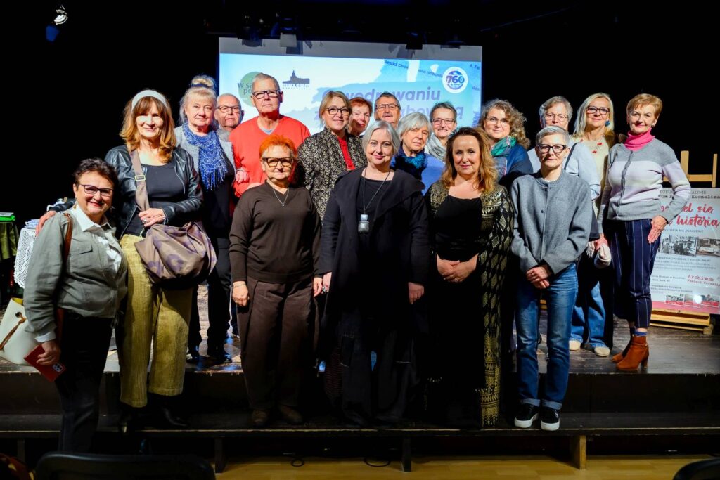 Grupa kilkunastu kobiet i mężczyzn pozuje do wspólnego zdjęcia na scenie; za uczestnikami widoczny jest ekran prezentacji z grafiką i logo Muzeum w Koszalinie oraz plansza wydarzenia (kliknięcie powiększy zdjęcie).