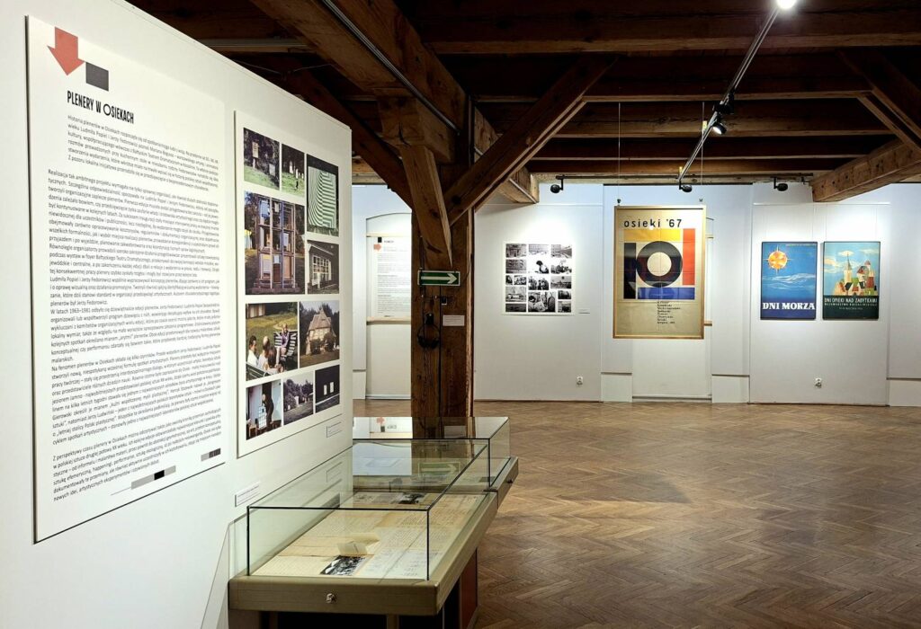 Sala wystawowa z drewnianym stropem i parkietem; na ścianach plansze informacyjne, fotografie oraz plakaty, a na pierwszym planie szklane gabloty z dokumentami i materiałami archiwalnymi (kliknięcie powiększy zdjęcie).